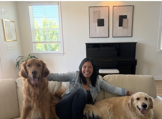 Two dogs and a latino woman sit on a tan couch in front of a piano