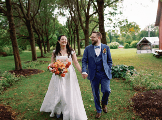 A bride in a white gown holding a vibrant orange and pink bouquet walks hand-in-hand and shares a laugh with her groom in a blue suit on a grassy path lined with trees.