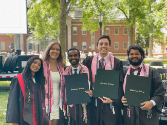 Five smiling graduates in caps and gowns, wearing pink stoles and holding Davidson College diploma covers, pose together outdoors on a campus lawn.
