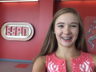 A young Courtney with braces smiles for a selfie in front of a red wall featuring a lighted ESPN logo.