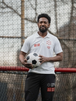 A young bearded man in a white polo holds a soccer ball