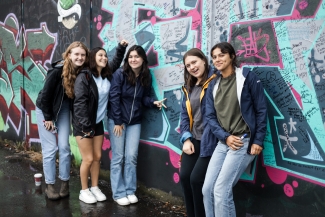 Five smiling young women pose together in front of a vibrant graffiti wall covered in written messages and colorful abstract shapes.