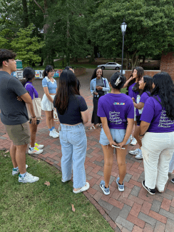 A diverse group of college students stands in a circle chatting on an outdoor brick path, with several students wearing purple t-shirts that read "Students Together Reaching Individually for Development and Education."