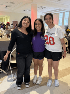 Three smiling young women pose together arm-in-arm in a well-lit banquet hall, with the student on the right wearing a white Davidson College t-shirt.