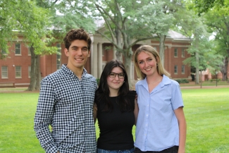 hree young adults smile and pose together arm-in-arm on a green grassy lawn with a brick college campus building and trees in the background