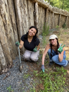 Two young women wearing gardening gloves crouch by a tall, rustic wooden log fence, smiling and flashing peace signs while weeding and using a small hand trowel in the dirt.