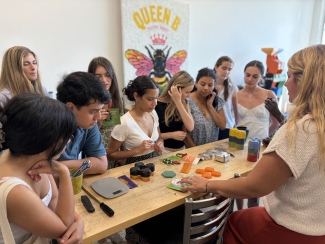 A group of young adults gathers around a wooden workshop table, listening intently as an instructor points to small, colorful, recycled plastic objects next to tools and a digital scale, beneath a "Queen B" bee poster.