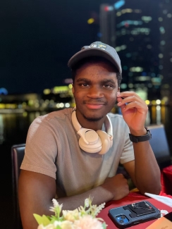A young man wearing a baseball cap and headphones around his neck smiles while seated at an outdoor waterfront restaurant at night.