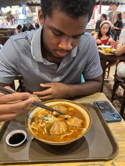 A young man uses chopsticks to eat a large bowl of noodle soup with meat pieces at a crowded restaurant table.