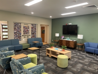 A spacious student lounge featuring blue and teal seating, light green accent walls, and a large photo wall titled "CSDI MEMORIES 25-26" next to a television screen.