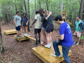 A group of students participates in an outdoor team-building exercise in a wooded area, carefully helping each other balance and walk across narrow wooden planks connected to low platforms.