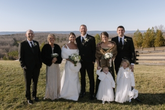 A wedding party poses together outdoors on a grassy hill with a scenic valley landscape in the background, featuring the bride in a white gown and fur wrap alongside the groom, bridesmaids in wraps, men in black suits, and two young flower girls in white dresses.