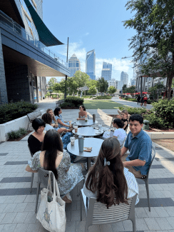 A group of students sits and chats around outdoor patio tables in a modern city plaza courtyard, with a grassy lawn, trees, and tall skyscrapers visible in the background under a clear blue sky.