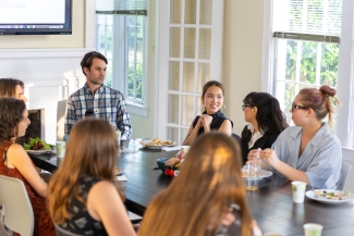 A group of young adults sits around a large conference table in a bright room with windows, enjoying a casual meal while listening to a woman speak during a group discussion.