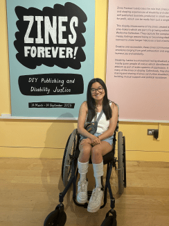 A smiling young woman using a wheelchair poses in front of a museum exhibit poster titled 'Zines Forever! DIY Publishing and Disability Justice'