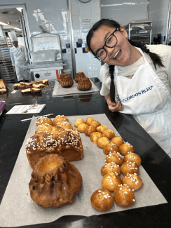 A smiling young woman wearing a Le Cordon Bleu apron leans over a bakery counter filled with freshly baked pastries, including brioche with pearl sugar and kouglof