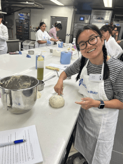 A smiling young woman wearing a Le Cordon Bleu apron shapes a round ball of dough on a commercial kitchen counter next to a metal mixing bowl, while other students work in the background