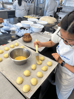 A student wearing a Le Cordon Bleu apron uses a pastry brush to apply an egg wash to small, round portions of dough arranged on a lined baking sheet.
