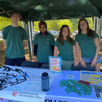 Four smiling students wearing green shirts stand behind a Davidson College Sustainability Office information table featuring an Earth Month poster and promotional buttons.