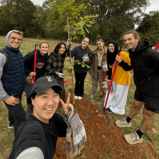 A diverse group of eight students smiles outdoors around a newly planted young oak tree, holding shovels and rakes in a grassy field.