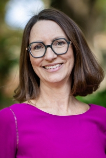 A woman with shoulder-length brown hair and glasses and a bright purple shirt