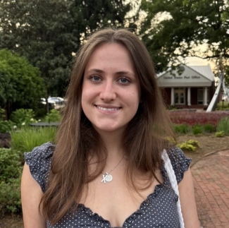 A smiling young woman with long brown hair, wearing a blue and white polka dot top and a necklace, stands on a brick path outdoors with a United States Post Office building in the background.