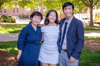 A young woman standing between two older adults (presumably her parents) on a sunny campus setting with brick buildings and trees in the background.