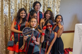 A group of cheerful graduates wearing ASAPI graduation stoles pose with playful props, including a sign that reads "Yep, they let me graduate," in front of a sparkly gold sequin backdrop.