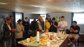 A group of people gathers around a kitchen island covered with food and drinks, watching and clapping as an older man cuts a cake.
