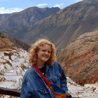 A smiling young woman with curly blonde hair, wearing a denim jacket and a colorful cross-body bag, stands in front of a mountain valley with stepped salt evaporation ponds in the background.