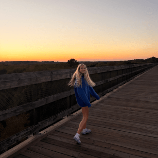 A smiling young woman with long blonde hair turns back toward the camera while walking along a wooden trestle bridge at sunset.