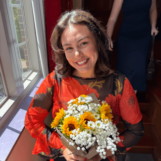 A smiling young woman with brown hair, wearing a long-sleeved red and black floral top, holds a bouquet of sunflowers and baby's breath near a sunny window.