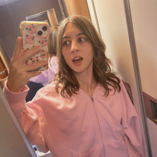 A young woman with shoulder-length light brown hair takes a mirror selfie wearing a light pink zip-up hoodie, holding a phone with a patterned case.
