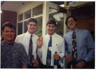 Four young men in dress shirts and ties pose together outside a building, with three of them holding bottles of champagne and smiling.