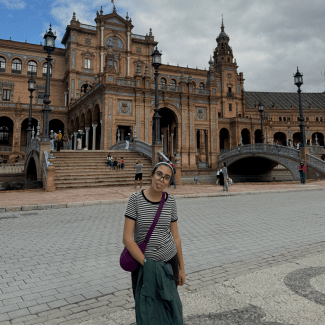 A young woman with glasses sticking her tongue out playfully stands in a paved plaza in front of the ornate, historic Plaza de España architecture and bridges in Seville.