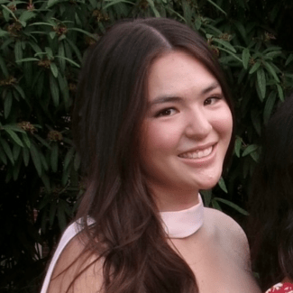 A smiling young woman with long dark brown hair, wearing a light pink dress with a high collar, poses outdoors in front of leafy green foliage.