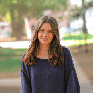 A smiling young woman with long brown hair, wearing a dark navy blue knit sweater, poses outdoors against a blurred, sunlit background.