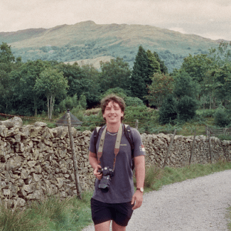 A smiling young man with curly brown hair walks down a gravel path carrying a camera around his neck, flanked by a stone wall with green hills and mountains in the background.