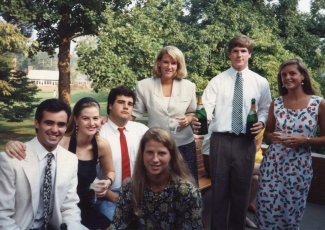 A vintage outdoor group photo of young adults dressed in semi-formal clothing, with two individuals holding green bottles of champagne.
