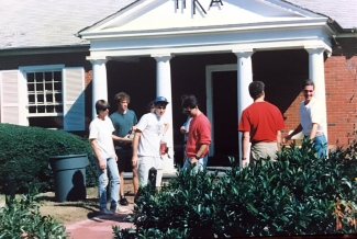 A group of young men in casual attire socialize outside a brick fraternity house with white columns under a sign with Greek letters.