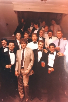 A group of young men posing together on a staircase while dressed in formal tuxedos and suits, some holding plastic cups.