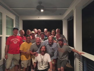 A large group of smiling middle-aged men pose closely together for a group photo on a porch at night.