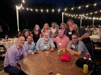 A group of men and women with name tags smile around a circular wooden table at an outdoor evening event lit by string lights.