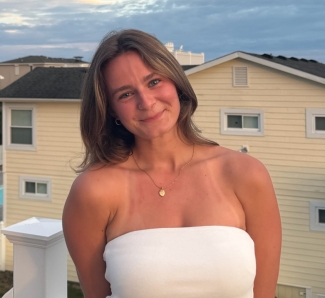 A smiling young woman with light brown hair and a nose ring, wearing a white tube top and a gold pendant necklace, stands outdoors with yellow residential houses and a cloudy sky in the background.