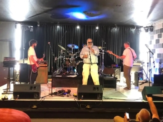 A live four-piece rock band with a lead singer in sunglasses performs on a brightly lit indoor stage with a black curtain backdrop.