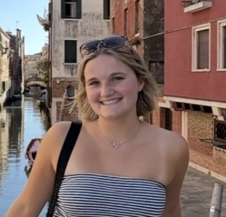 A smiling young woman with blonde hair, wearing sunglasses on her head and a striped tube top, poses on a bridge overlooking a canal in Venice.