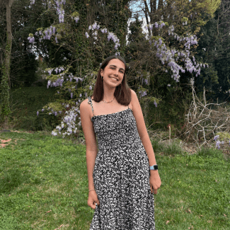 A smiling young woman with shoulder-length dark hair, wearing a black and white patterned sundress, stands on a grassy lawn in front of blooming purple wisteria trees.