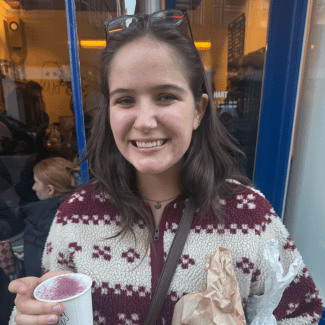 A smiling young woman with brown hair, glasses resting on top of her head, and a nose ring wears a patterned fleece jacket while holding a beverage with purple foam and a paper bag outside a storefront.