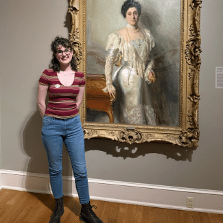 A smiling young woman with dark curly hair and glasses, wearing a striped scoop-neck shirt and blue jeans, stands inside an art gallery next to a large framed oil portrait of a woman in an elegant white gown.