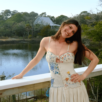 A smiling young woman with long dark hair, wearing a white floral-embroidered dress, tilts her head while leaning against a white railing overlooking a pond surrounded by lush trees and a house.